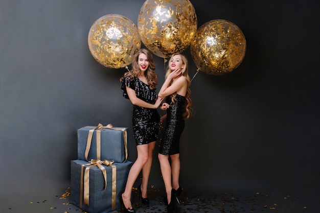 Free photo happy party time of two charming young women in luxury black dresses. long curly hair, attractive look, presents, big balloons with golden tinsels, smiling, having fun.