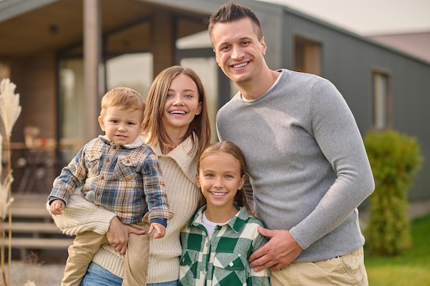 Happy people.. Young beautiful woman with little boy attractive man and long-haired school-age girl in casual clothes hugging joyful looking at camera standing near house