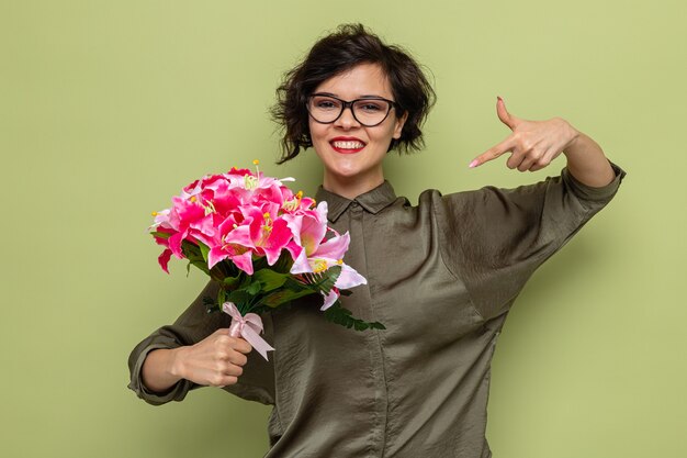 Happy and pleased woman with short hair holding bouquet of flowers pointing with index finger at it smiling cheerfully celebrating international women's day march 8 standing over green background