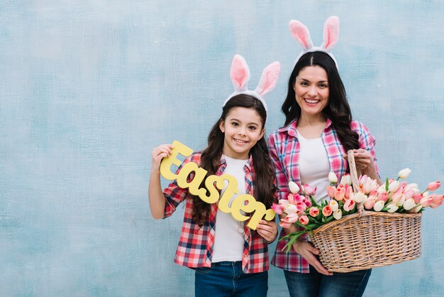 Happy portrait of mother and daughter holding easter word and tulips basket against blue wall