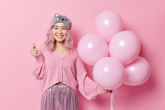 Happy positive Asian woman makes korean like sign shapes mini heart expresses love holds bunch of balloons wears sleepmask jumper and skirt celebratess birthday isolated over pink background