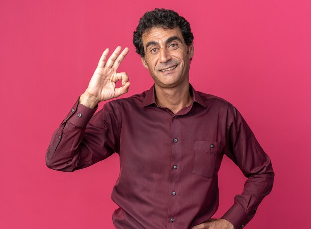 Free Photo happy and positive senior man in purple shirt looking at camera smiling cheerfully doing ok sign standing over pink background