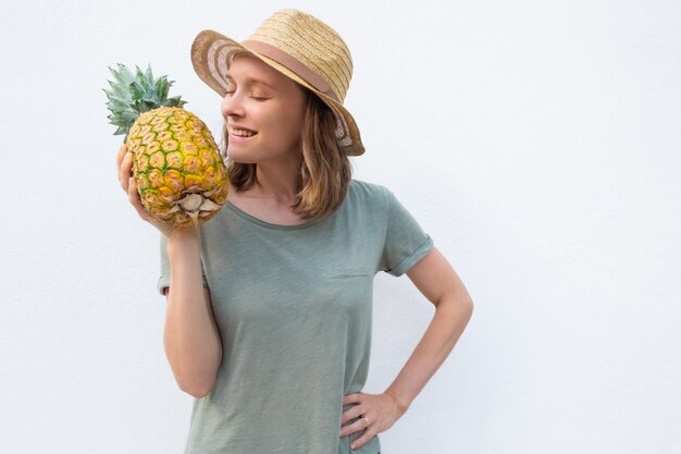 Happy positive woman in summer hat smelling whole pineapple