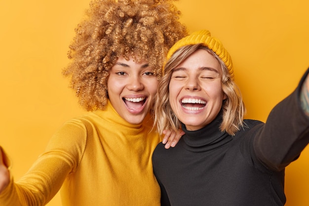 Happy positive young women have fun laugh gladfully take selfie dressed in casual clothes stand next to each other isolated over yellow background Cheerful female friends photograph themselves