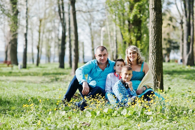 Happy pregnant family with two sons dressed in a turquoise clothes sitting on grass with flowers at park