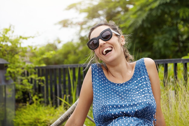 Happy pregnant woman wearing sunglasses and blue dress relaxing outdoors against green nature