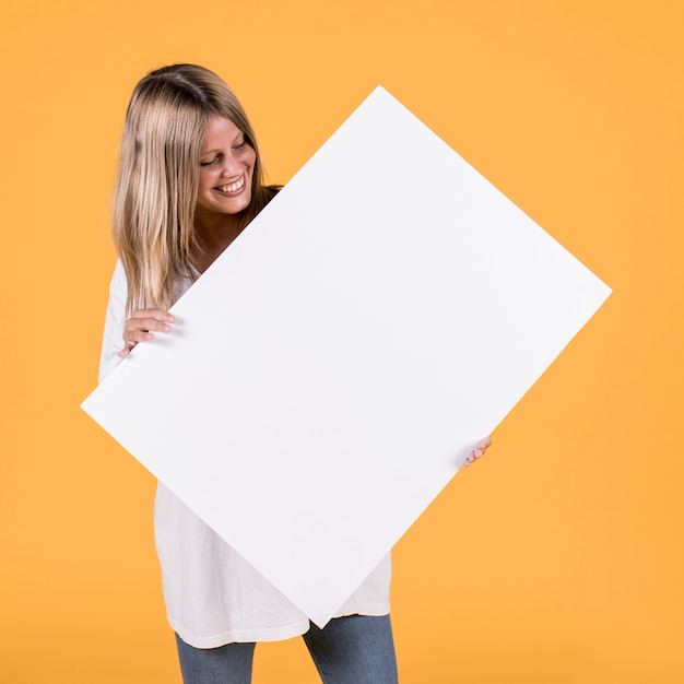 Happy pretty woman holding blank white cardboard against yellow wallpaper