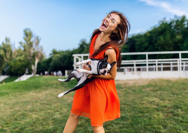 Happy pretty woman park holding boston terrier dog, smiling positive mood, trendy summer style, wearing orange dress, sunglasses, playing with pet, having fun, colorful