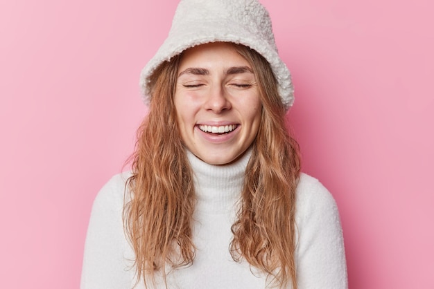 Free Photo happy pretty woman with long hair keeps eyes closed smiles joyfully has dimples on cheeks expresses positive authentic emotions wears panama and white warm sweater isolated over pink background.