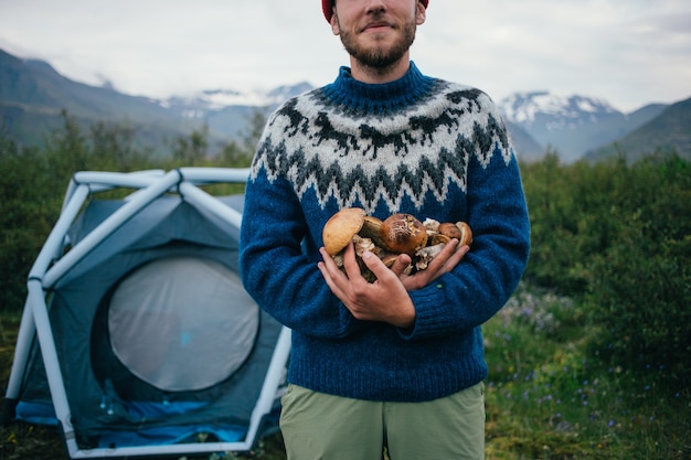 Happy, proud picker man in traditional blue wool sweater with ornaments stands on camping ground in mountains, holds in arms pile of delicious and organic mushrooms