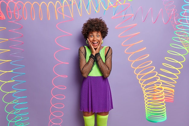 Happy reaction concept. Joyful dark skinned woman keeps hands on cheeks, stares with overjoyed expression, wears colorful outfit