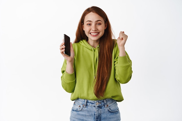 Happy redhead girl rejoice, holding smartphone and celebrating, winning on mobile phone and triumphing, purchase with discount, standing on white