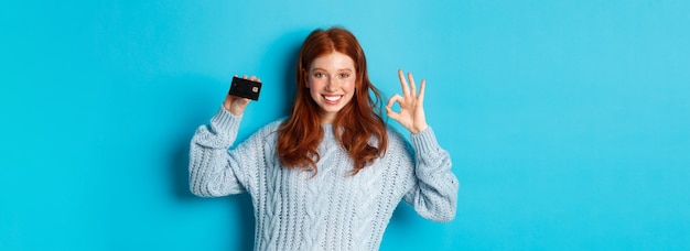 Free photo happy redhead girl in sweater showing credit card and okay sign recommending bank offer standing ove