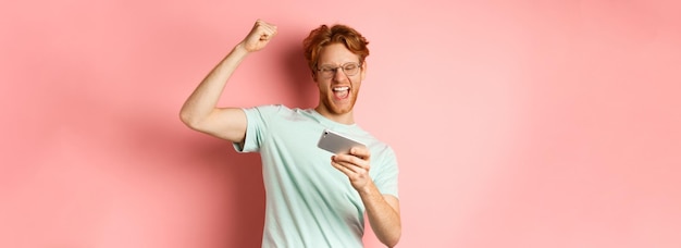 Free photo happy redhead man winning in mobile video game raising hand up and shouting yes with joy celebrating