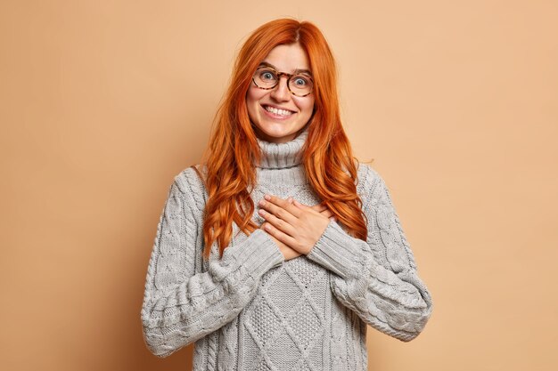 Happy redhead woman keeps hands to chest makes gratitude gesture smiles happily wears knitted grey sweater.