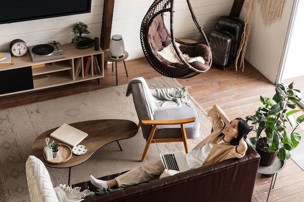 Happy relaxed asian woman laying on couch at home with laptop listening podcast and enjoying weekend