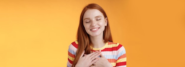 Free photo happy romantic delighted sighing young lovely redhead girlfriend smiling broadly touching heart chest palms close eyes recalling date passionate kiss feel gratitude warmth orange background