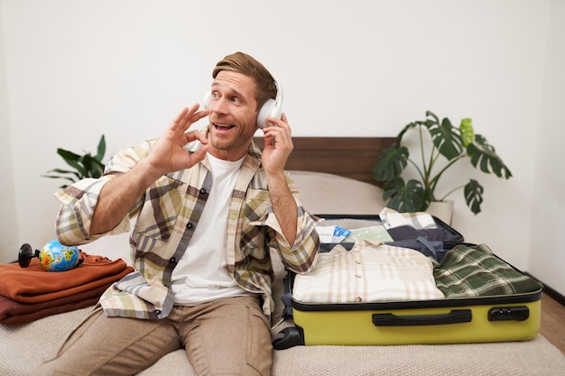 Free photo happy satisfied young man shows approval gesture sits with wireless headphones packs luggage tourist