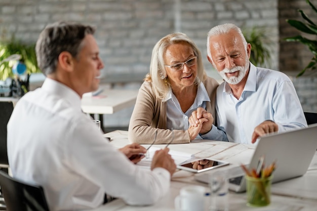 Happy senior couple holding hands and using laptop while having a meeting with financial advisor in the office Senior man is pointing at something on laptop