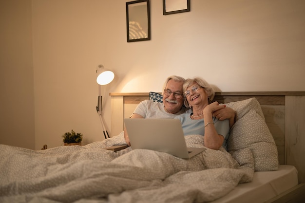 Happy senior couple lying down in bed and surfing the net on laptop at night