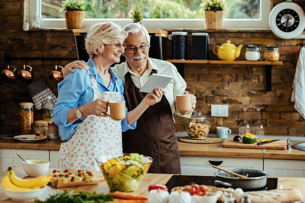 Happy senior couple surfing the net on touchpad while drinking coffee in the kitchen.