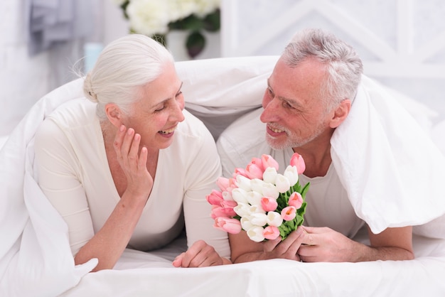 Happy senior woman lying on bed looking at tulip flowers held by her husband