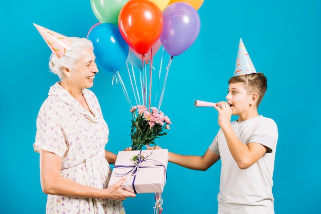 Free Photo happy senior woman with birthday gift and flowers looking at her grandson blowing party horn