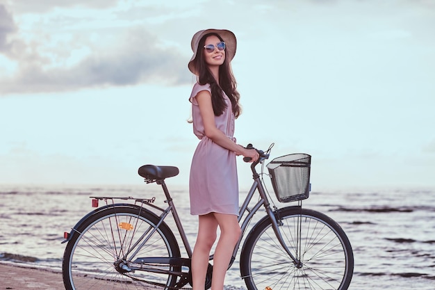 Happy sensual girl in sunglasses and hat wearing dress walks with her bicycle on the beach against amazing seaside background.