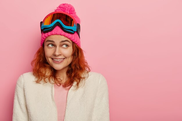 Happy skier girl smiles gently, focused aside, wears pink winter hat with pompon, white soft sweater on zip, ski glasses on head, poses against rosy wall