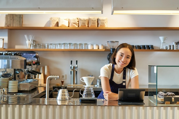 Free photo happy smiling asian barista girl behind counter working with pos terminal and brewing filter kit mak