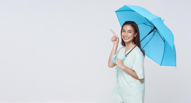 Happy smiling Asian woman standing and holding blue umbrella and pointing finger to copy space