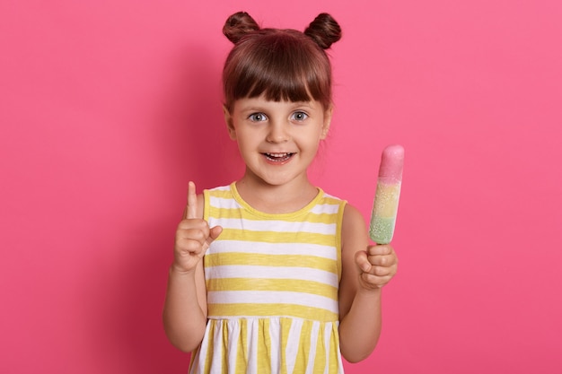 Happy smiling kid girl holding ice cream and pointing up with her index finger, wearing summer dress with white and yellow stripes.