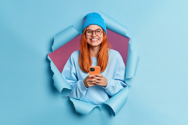 Free photo happy smiling millennial girl with red hair holds modern cellular enjoys texting in social media uses mobile network services wears blue jumper and hat.