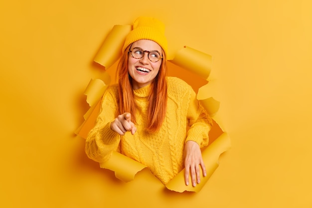 Happy smiling redhead woman points towards, wears hat sweater and spectacles.