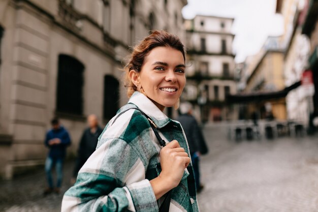 Happy smiling woman with blond hair wearing striped shirt walking on the sunny old street. Young girl with bag is walking in the city