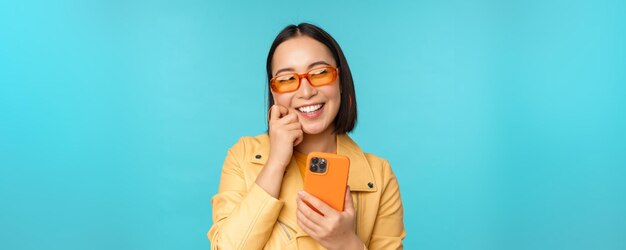 Happy stylish asian girl using smartphone and laughing smiling at camera standing over blue background