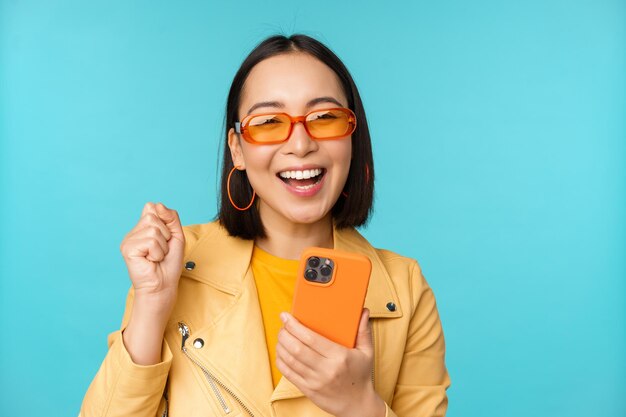 Happy stylish asian girl using smartphone and laughing smiling at camera standing over blue background