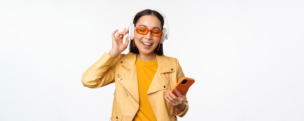 Happy stylish asian woman in sunglasses listening music in headphones holding smartphone singing and dancing standing over white background