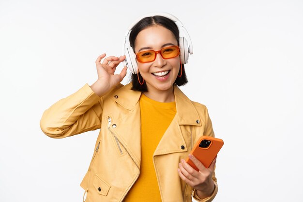 Happy stylish asian woman in sunglasses listening music in headphones holding smartphone singing and dancing standing over white background