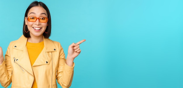 Happy stylish chinese girl in sunglasses points fingers sideways invites to check out see store discount shows left and right stands over blue background