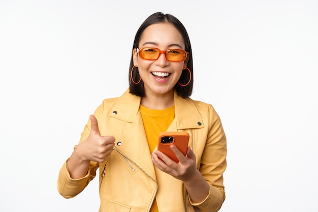 Happy stylish korean girl wearing sunglasses showing thumb up holding smartphone using mobile phone app recommending application online store white background