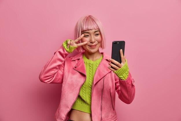 Happy stylish millennial girl shows two fingers over eye, peace sign, takes selfie, enjoys her new hairstyle, dyed hair in pink