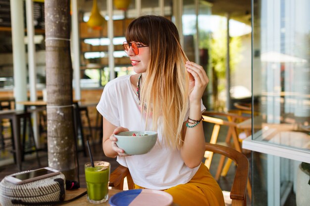 Happy stylish woman eating healthy food sitting in the beautiful interior with green flowers