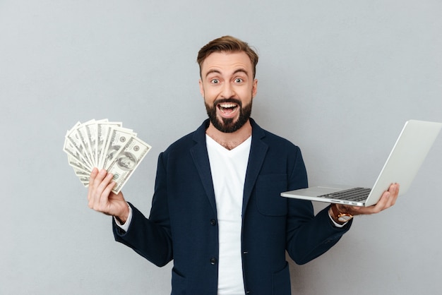 Happy surprised bearded man in busines clothes holding money and laptop computer while looking at the camera over gray 