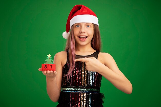 Happy and surprised little girl in glitter party dress and santa hat showing toy cubes with new year date pointing with index finger at it smiling cheerfully standing over green background