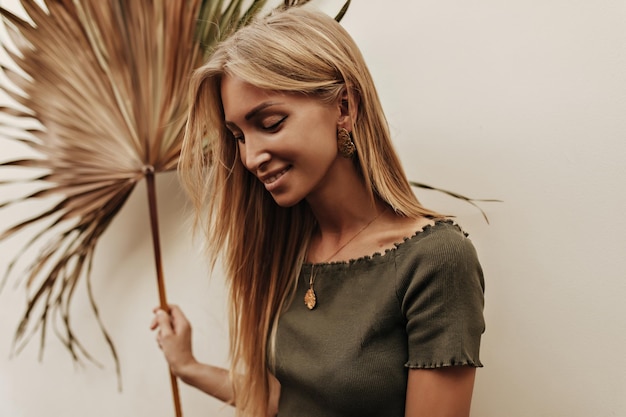 Free photo happy tanned woman in dark green tshirt smiles sincerely and poses outside blonde girl in khaki top holds palm leaf on white wall background