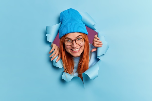 Free photo happy teenage girl breaks through paper wall has fun looks gladfully wears blue hat spectacles smiles toothily