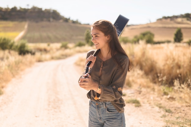 Free photo happy teenage girl standing on dirt road holding guitar