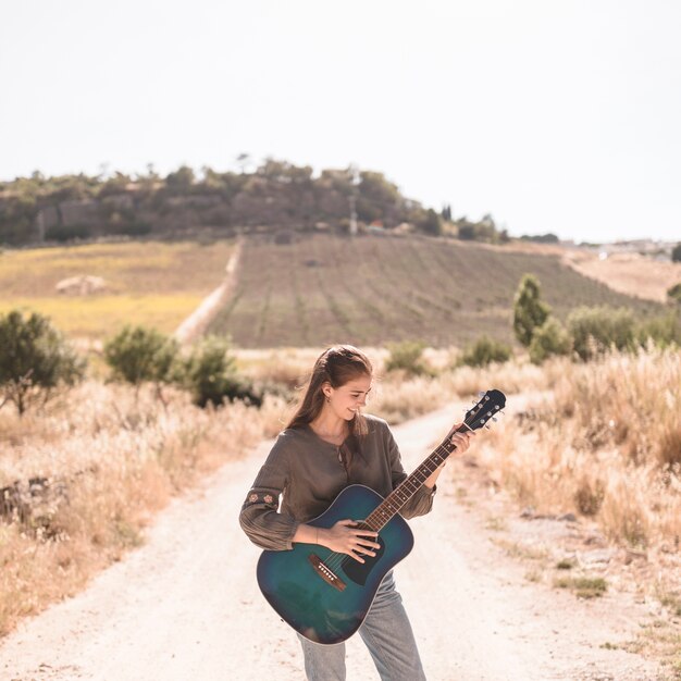 Happy teenage girl standing on dirt track playing guitar