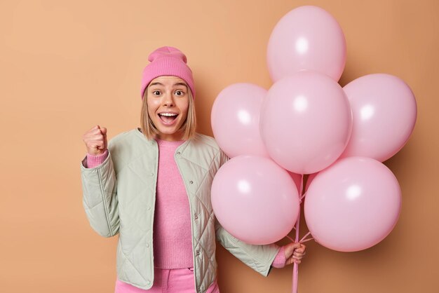 Free photo happy teenage girl with fair hair clenches fist celebrates good news dressed in jacket and pink hat holds bunch of inflated balloons celebrates special occasion isolated over brown background.